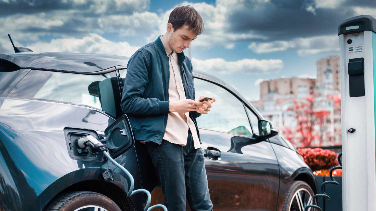 Mężczyzna opiera się o swój samochód elektryczny podczas jego ładowania i spokojnie przygląda swój telefon, ponieważ nie martwi się o nowy podatek od samochodów spalinowych dla przedsiębiorców w Holandii, z którego elektryki są zwolnione.